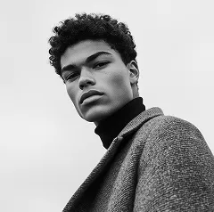 Black and white portrait of a young man with curly hair wearing a turtleneck and coat looking confidently at the camera.